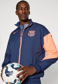 Marineblå og orange Nike jakke med lynlås foran, stående krave, holdningslogo og sidelommer; modellen holder en hvid fodbold.