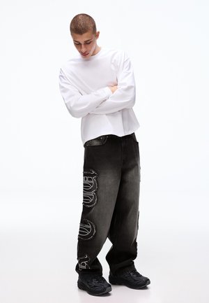 Nuori mies päällään väljät mustat farkut valkoisilla laikuilla, valkoinen pitkähihainen paita ja mustat lenkkarit, seisoo käsivarret ristissä, katsoen alas.