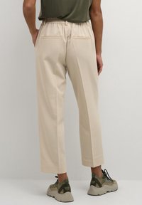 Pantaloni dritti beige con vita elastica, due tasche posteriori e una texture liscia, abbinati a sneakers con lacci a fantasia.