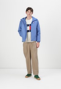 Jachetă cu glugă de culoare albastru deschis, cu manșete elastice, purtată peste un pulover crem cu un design de logo în nuanțe de navy și roșu. Pantaloni bej și pantofi verzi.