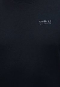 Μαύρο ύφασμα με μικρό λευκό κείμενο που γράφει "1962" και "Tom Tailor" στην επάνω αριστερή περιοχή του στήθους.