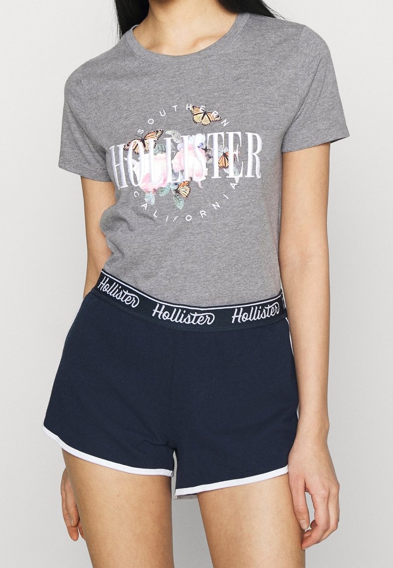 Grå kortärmad t-shirt med blommigt och fjärilsdesign; kombinerad med marinblå shorts med midjeband i vitt.