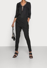 Schwarze Bluse mit V-Ausschnitt und dekorativen Verzierungen, kombiniert mit schwarzen, zulaufenden Hosen und schwarzen Sandaletten mit Absatz. Accessoires umfassen eine kleine schwarze Handtasche.