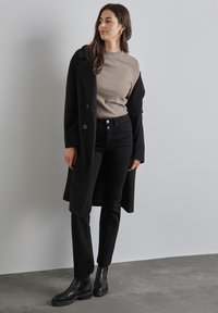 Zwarte jas, beige geribbelde top en zwarte jeans. De outfit heeft een gladde textuur, een getailleerde pasvorm en zwarte enkellaarsjes.