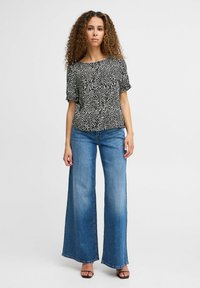 Camicia a maniche corte con motivo in bianco e nero abbinata a jeans a gamba larga azzurri e sandali neri con tacco. Sfondo semplice.