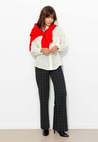 Chemise blanche en broderie anglaise avec un col arrondi, associée à un pantalon large à carreaux noirs et un pull rouge noué aux épaules. Talons noirs.