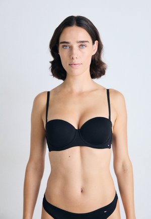 Jonge vrouw met kort bruin haar, gekleed in een zwarte strapless beha en bijpassend zwart ondergoed tegen een effen lichte achtergrond.