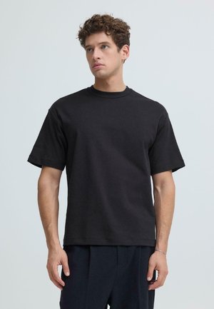 Ung mand med krøllet hår iført en simpel sort kortærmet T-shirt og mørke bukser, stående mod en neutral baggrund.