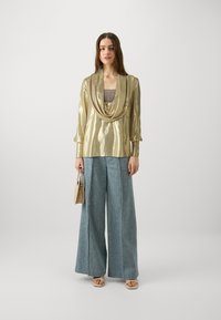Top metallico oro con scollatura drappeggiata, abbinato a jeans a gamba larga blu chiara. La modella tiene una borsa beige. Sandali semplici completano il look.