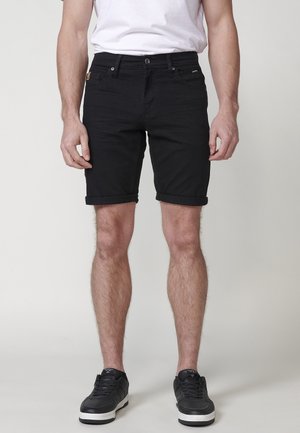 Man wearing black rolled-hem shorts, white t-shirt, and black sneakers, standing against a plain light background.