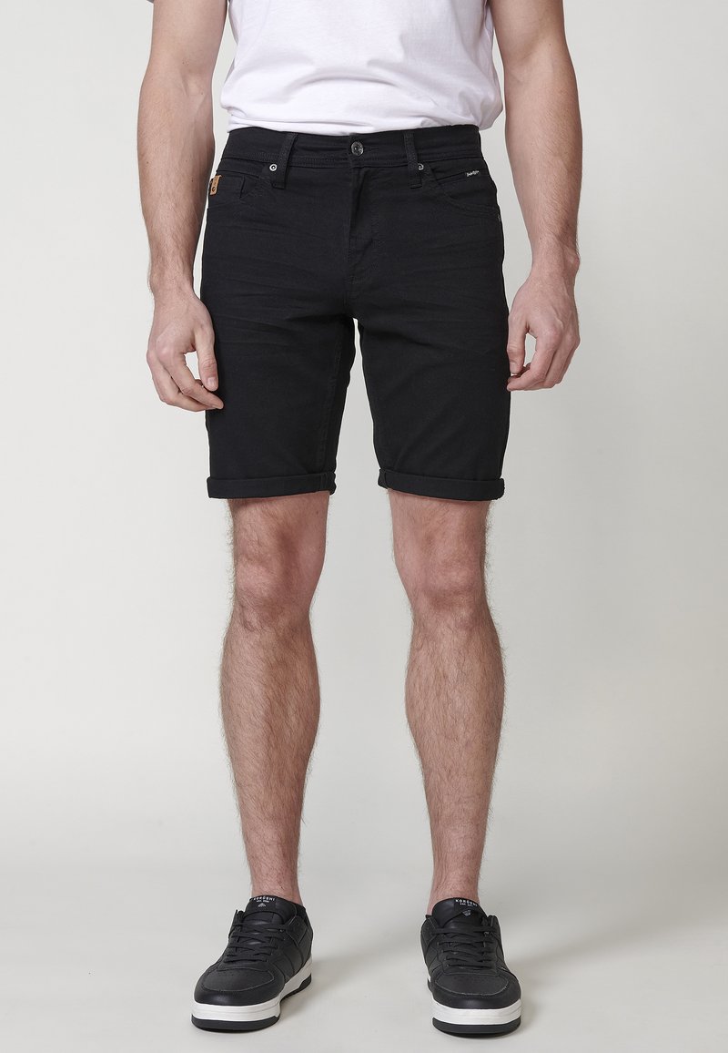 Mies, joka on pukeutunut mustiin kääntöhelmanshortseihin, valkoiseen t-paitaan ja mustiin lenkkareihin, seisoo yksinkertaista vaaleaa taustaa vasten.