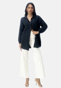 Camicia nera a maniche lunghe con texture e bottoni, abbinata a pantaloni bianchi a gamba larga e sandali neutri. Design semplice, silhouette fluida.