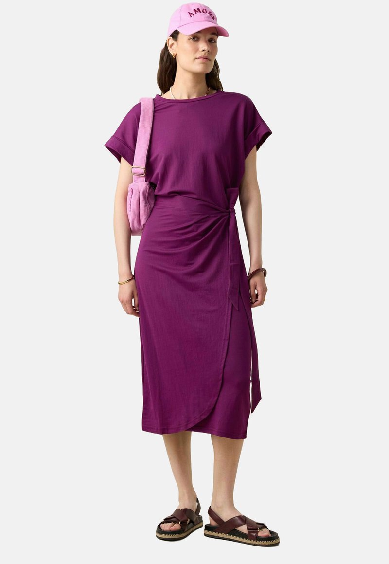 Robe portefeuille violette à manches courtes, avec détail de nœud sur le côté ; assortie à une casquette rose et un sac à épaule, ainsi qu'à des sandales à lanières marron.