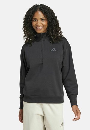 Mujer sonriente con cabello rizado lleva un sudadera negra con cremallera media y pantalones blancos, de pie contra un fondo claro y liso.
