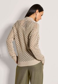 Vrouw met lang, donker haar draagt een beige opengehaakt trui en olijfgroene textuurbroek, poserend met handen in haar zakken tegen een effen achtergrond.