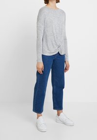 Haut gris à manches longues avec un détail noué à l'avant, associé à un jean bleu coupe droite et des baskets blanches. Texture lisse et coupe décontractée.