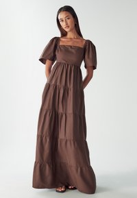 Brown cotton maxi dress with puff sleeves, square neckline, and tiered design. Smooth texture and ankle-length hemline.