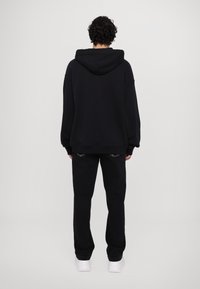 Felpa nera oversize con cappuccio, caratterizzata da una vestibilità ampia e polsini a coste, abbinata a pantaloni neri e sneakers bianche.
