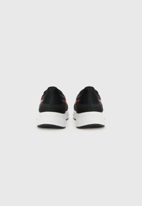 Baskets noires de sport avec un dessus en mesh, accents rouges, une semelle intermédiaire blanche et des sections de talon noires texturées. Design minimaliste avec un bout arrondi.