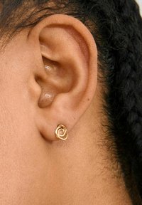 Gouden ronde studoorbel met een gestructureerd, gedraaid ontwerp. Draagt in de oorlel, naast een close-up van een huidtint en gevlochten haar.