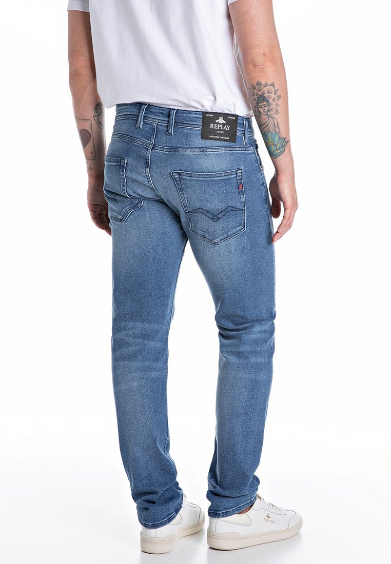 Blaue Denim-Jeans mit slim fit, mit Gesäßtaschen und einem schwarzen Etikett mit "REPLAY". Kombiniert mit einem schlichten weißen Shirt und weißen Sneakers.