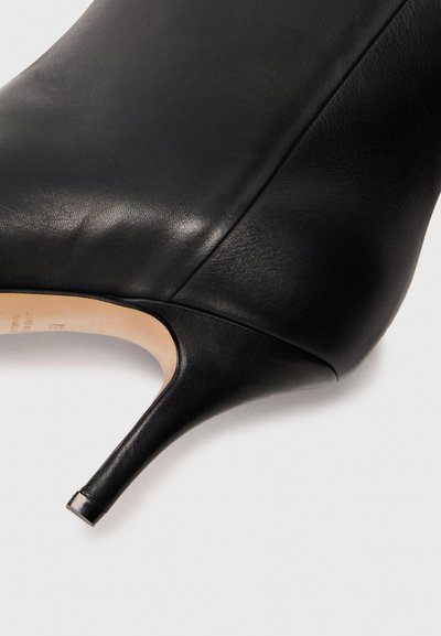 Chaussure à talon haut en cuir noir avec un bout pointu et un talon fin et courbé. La texture est lisse avec de légères rides et une finition mate.