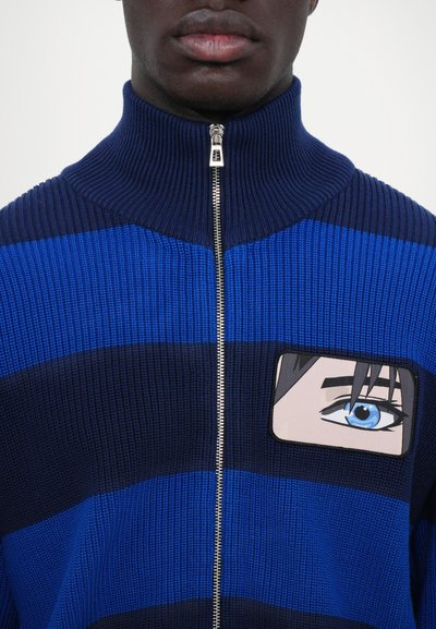Blue and navy striped zip-up sweater with ribbed texture, featuring an embroidered patch of a face with a blue eye on the chest.