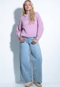 Jasnoróżowy sweter z dzianiny z prążkowanym dołem, szerokie jasnoniebieskie dżinsy oraz beżowe trampki na platformie, prezentowane na modelce na białym tle.