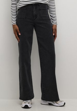 Person iført sorte vidde jeans, hvid og sort stribet langærmet skjorte og hvide sneakers, der står mod en ensfarvet baggrund.