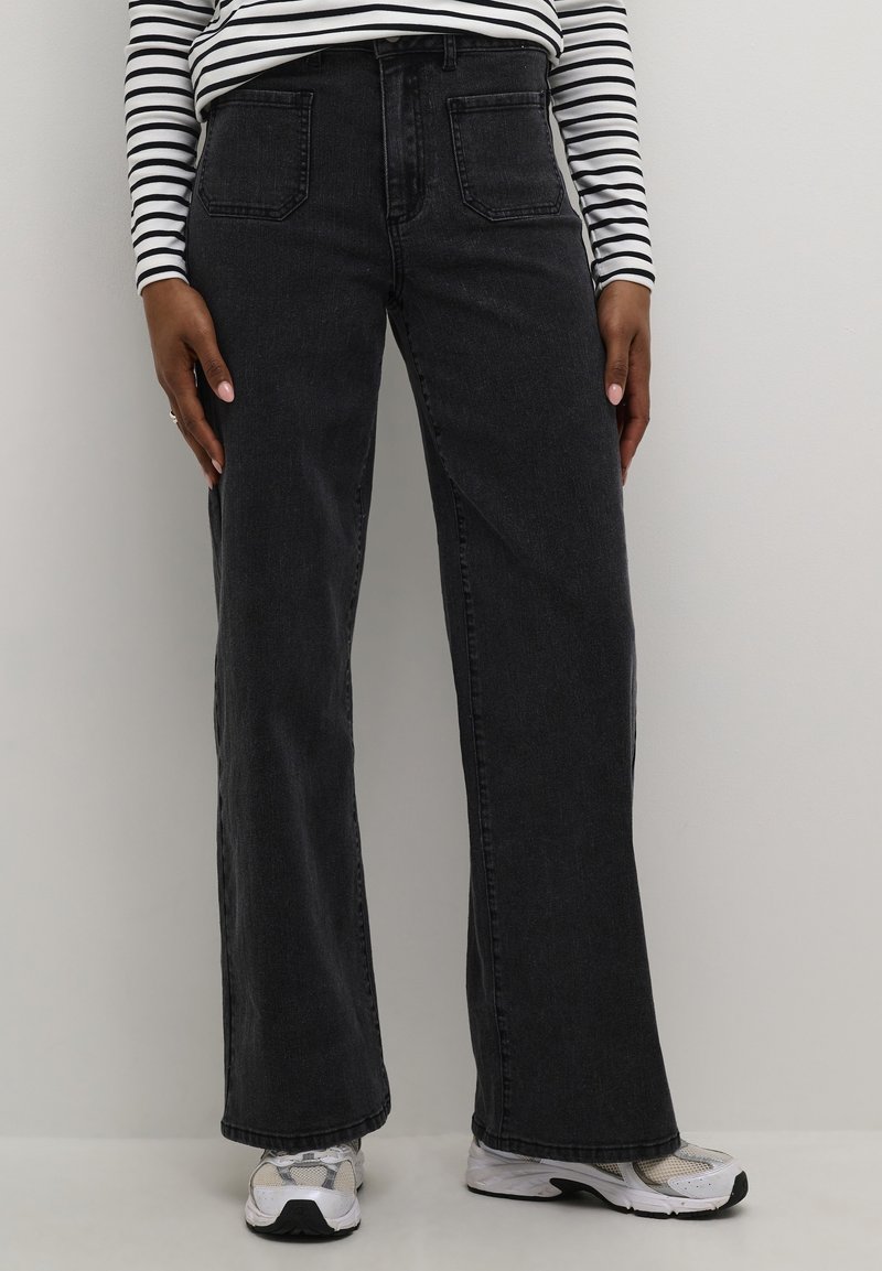 Personne portant un jean noir à jambes larges, un t-shirt manches longues rayé blanc et noir, et des baskets blanches, debout devant un fond uni.