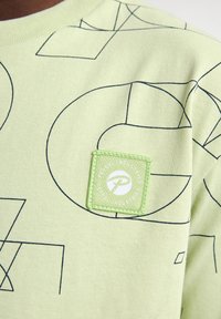 Lichtgroene katoenen t-shirt met een geometrisch patroon in zwarte lijnen, met een vierkante groene patch met een logo aan de linkerkant.