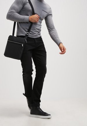Sac bandoulière noir avec un design texturé, porté avec une chemise grise à manches longues et un pantalon noir, complété par des baskets noires.