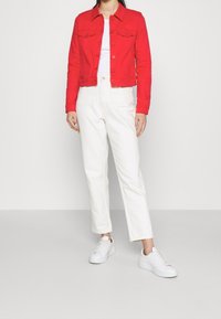 Chaqueta de mezclilla roja con dos bolsillos en el pecho, camiseta blanca debajo y pantalones blancos de cintura alta. El modelo lleva zapatillas blancas.