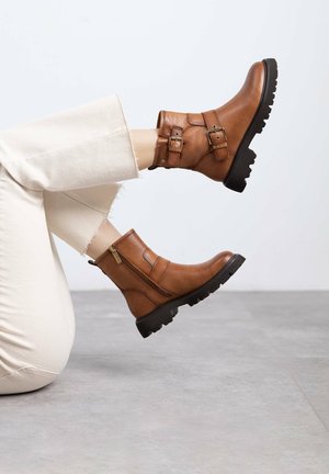 Botas de cuero marrón hasta el tobillo con cremallera lateral, doble hebilla, suelas de goma negra gruesa y superficie texturizada. Combinadas con pantalones cortos de color claro.
