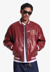 Bărbat cu păr creț și mustață, purtând ochelari de soare negri, jachetă varsity de piele roșie cu dungi albe, cămașă albă și pantaloni închiși la culoare.