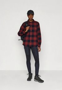 Camisa de flanela vermelha e preta com padrão xadrez, leggings pretas e botas de caminhada pretas. Os acessórios incluem um boné preto e uma mochila preta.