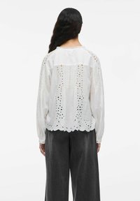 Frau mit langen dunklen Haaren, die eine weiße bestickte Bluse mit langen Ärmeln und lockeren schwarzen Hosen trägt, steht mit dem Rücken zu einer weißen Hintergrund.