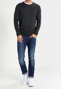 Dunkelgrauer Strickpullover mit Rundhalsausschnitt und weißem Innenbesatz, kombiniert mit blauen Jeans und weißen Sneakern. Der Pullover hat ein kleines Logo.
