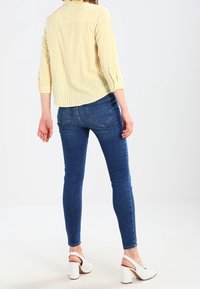 Yellow striped blouse, three-quarter sleeves, paired with dark blue skinny jeans and white block-heeled shoes, viewed from the back.