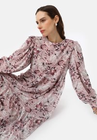 Robe florale en rose doux, avec des manches longues bouffantes et un col rond. Tissu fluide avec des motifs délicats dans des teintes de bourgogne et de crème.