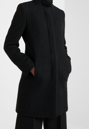 Schwarzer Textilwollmantel mit verdecktem Frontverschluss, hohem Kragen und Händen in den Seiten Taschen über schwarzen Hosen.