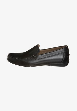 Μαύρα δερμάτινα loafers χωρίς δέσιμο με λείο φινίρισμα, ραφές κατά μήκος του άνω μέρους και στρογγυλή μύτη, τοποθετημένα σε μια επίπεδη καουτσούκ σόλα.