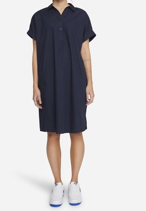 Persona che indossa un vestito camicia largo blu navy a maniche corte e sneakers bianche con suola blu, in piedi davanti a uno sfondo semplice.