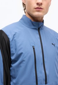 Άνδρας που φοράει μπλε και μαύρο μπουφάν με φερμουάρ και ψηλό γιακά, με λογότυπο Puma στο στήθος.