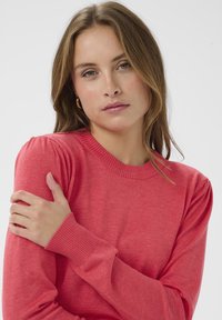 Pull-over rouge avec col et poignets côtelés, manches légèrement bouffantes et texture douce. Le tissu semble léger.