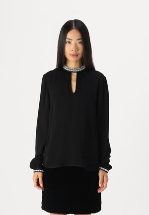 Black long-sleeve blouse with a keyhole neckline and embellished collar. Soft fabric and fitted cuffs with decorative accents. Matched with a black textured skirt.