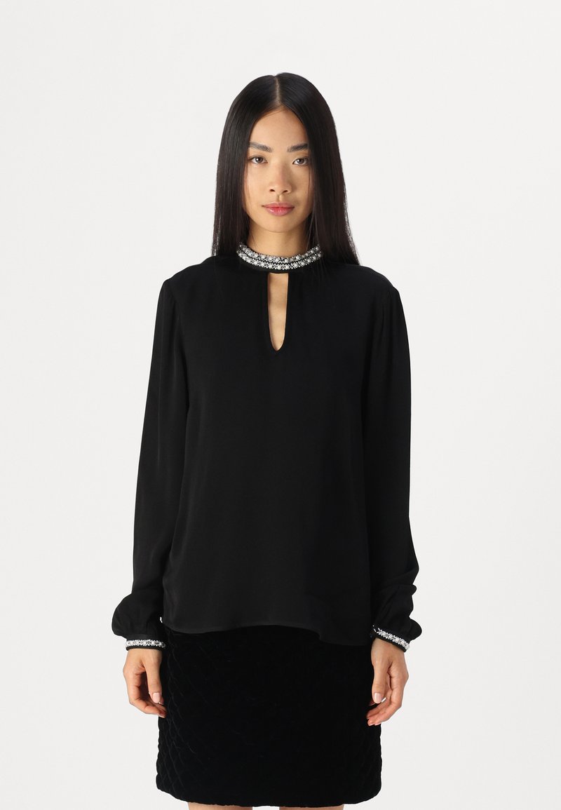 Blouse noire à manches longues avec un décolleté en trou de serrure et un col orné. Tissu doux et poignets ajustés avec des accents décoratifs. Associée à une jupe noire texturée.