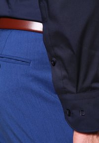 Navy-Button-Up-Hemd mit glatter Textur, schwarzem Manschettenaufschlag mit Knopf, kombiniert mit blaue, strukturierte Stoffhose und brauem Gürtel.