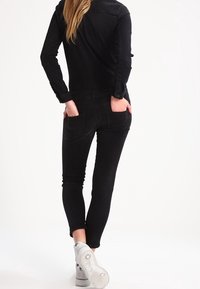 Černý denimový overal s přiléhavým střihem, dlouhými rukávy a zadními kapsami. V kombinaci s bílými teniskami, které podtrhují elegantní, moderní vzhled.