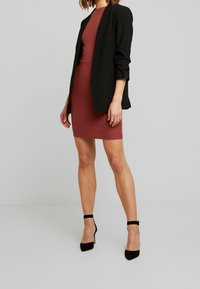 Blazer noir à manches longues porté sur une robe ajustée en maille côtelée couleur bordeaux. La robe arrive au genou et le mannequin porte des escarpins noirs à lanières.